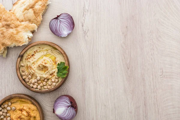 Vue du dessus de délicieux houmous aux pois chiches dans un bol près d'une pita fraîche cuite au four, oignon rouge sur table en bois beige — Photo de stock