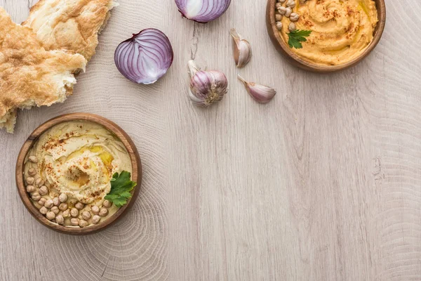 Vista dall'alto di delizioso hummus con ceci in ciotola vicino alla pita appena sfornata, aglio e cipolla rossa sul tavolo di legno beige — Foto stock