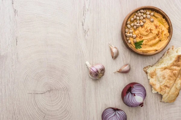 Vista superior do delicioso húmus com grão de bico na tigela perto de pita cozido no forno fresco, alho e cebola vermelha na mesa de madeira bege — Fotografia de Stock