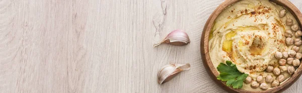 Vue du dessus de délicieux houmous aux pois chiches dans un bol près de l'ail sur une table en bois beige, vue panoramique — Photo de stock