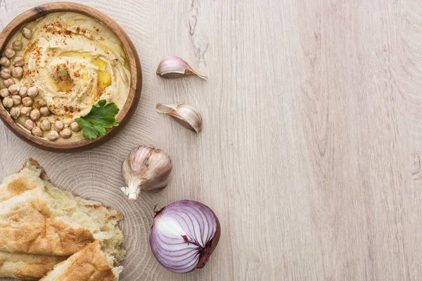 Vista dall'alto di delizioso hummus con ceci in ciotola vicino alla pita appena sfornata, aglio e cipolla rossa sul tavolo di legno beige — Foto stock