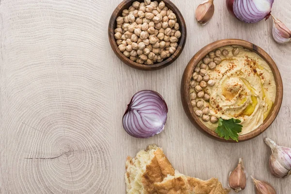 Blick von oben auf köstlichen Hummus mit Kichererbsen in Schüssel in der Nähe von frisch gebackener Pita, Knoblauch und roter Zwiebel auf beigem Holztisch — Stockfoto