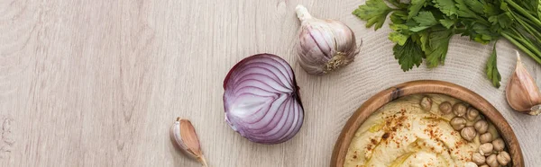 Vista dall'alto di delizioso hummus con ceci in ciotola vicino prezzemolo, aglio e cipolla rossa sul tavolo di legno beige, colpo panoramico — Foto stock