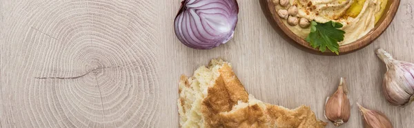 Draufsicht auf köstlichen Hummus mit Kichererbsen in Schüssel in der Nähe von frisch gebackenem Fladenbrot, Knoblauch und roter Zwiebel auf beigem Holztisch, Panoramaaufnahme — Stockfoto