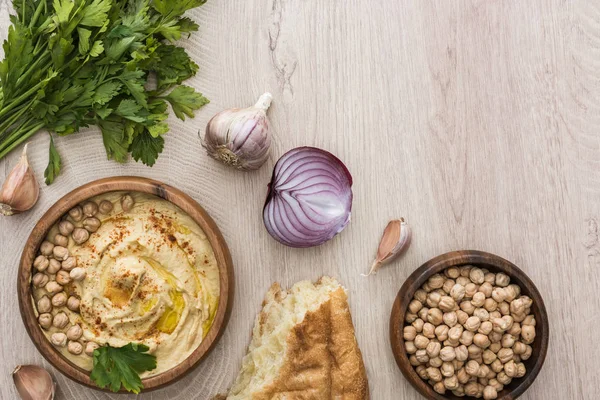 Vue de dessus de délicieux houmous aux pois chiches dans un bol près de pita fraîche cuite au four, ail, persil et oignon rouge sur table en bois beige — Photo de stock