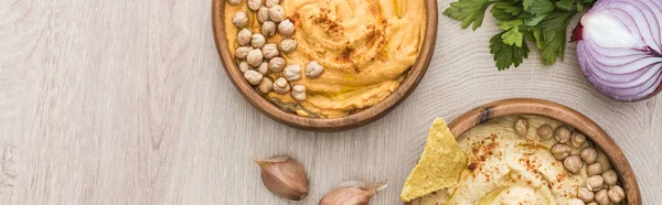 Top view of delicious hummus with chickpeas, nacho in bowls near spices and parsley on beige wooden table, panoramic shot — Stock Photo