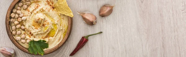 Vista dall'alto di delizioso hummus con ceci, nacho in ciotola vicino alle spezie e prezzemolo sul tavolo di legno beige, colpo panoramico — Foto stock