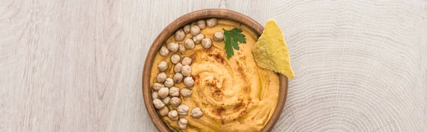 Draufsicht auf köstlichen Hummus mit Kichererbsen und Nacho in Schale auf beigem Holztisch, Panoramaaufnahme — Stockfoto