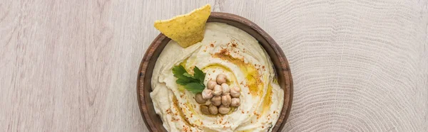 Vista dall'alto di delizioso hummus con ceci e nacho in ciotola su tavolo beige in legno, colpo panoramico — Foto stock