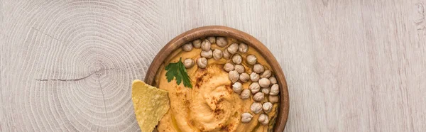 Vista superior de delicioso hummus com grão de bico e nacho na tigela em mesa de madeira bege, tiro panorâmico — Fotografia de Stock