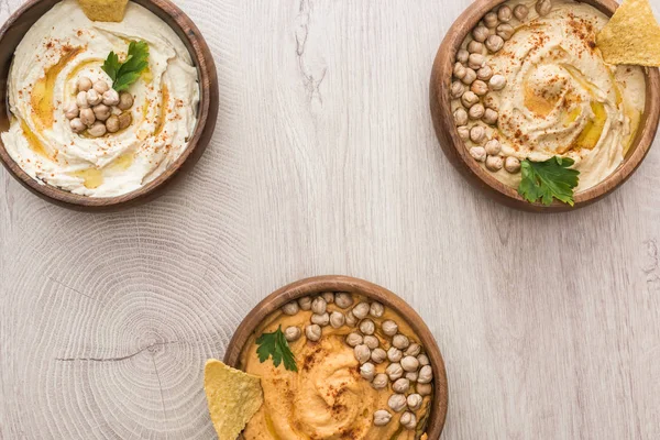 Vista superior del delicioso hummus con garbanzos y nachos en cuencos sobre mesa de madera beige - foto de stock