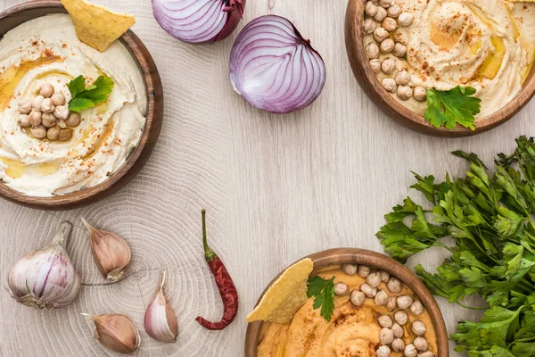 Vue de dessus de délicieux houmous aux pois chiches, nachos dans des bols près des épices et persil sur table en bois beige — Photo de stock