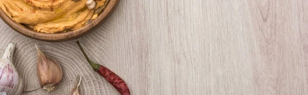 Top view of delicious hummus with chickpeas in bowl near garlic and chili pepper on beige wooden table, panoramic shot — Stock Photo