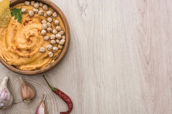 Vista superior do delicioso húmus com grão de bico, nacho na tigela perto de alho e pimenta na mesa de madeira bege — Fotografia de Stock