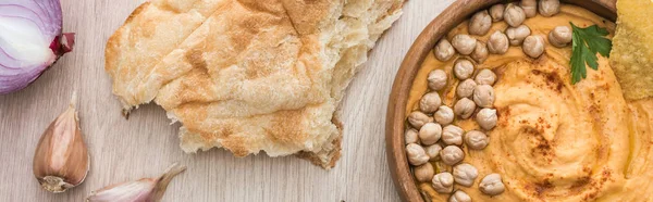 Vista superior de delicioso hummus com grão de bico em tigela perto de pita cozido no forno fresco e especiarias em mesa de madeira bege, tiro panorâmico — Fotografia de Stock