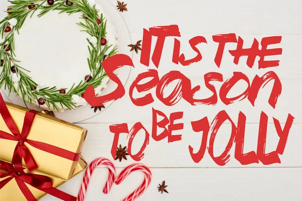 Top view of christmas pie, anise star seeds, candy canes and gift boxes on white wooden table with it is the season to be jolly lettering — Stock Photo