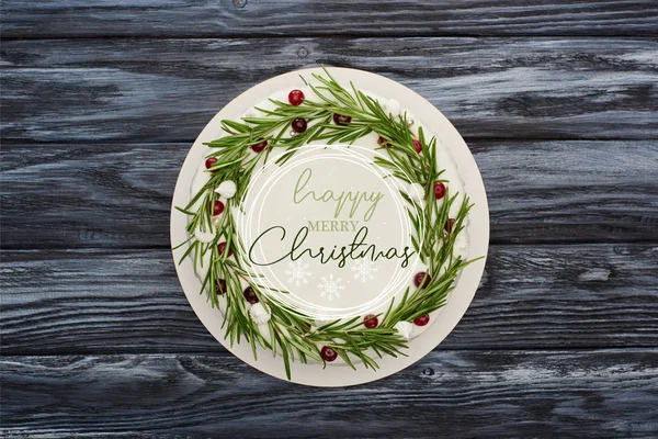 Vista superior del pastel de Navidad tradicional con glaseado blanco, romero y arándanos en la mesa de madera oscura con feliz ilustración de Feliz Navidad - foto de stock