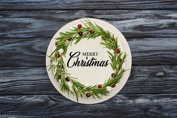 Vista superior del pastel de Navidad tradicional con glaseado blanco, romero y arándanos sobre una mesa de madera oscura con ilustración de Feliz Navidad - foto de stock