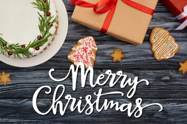Vista superior de pastel de Navidad, galletas y cajas de regalo en mesa de madera oscura con ilustración de Feliz Navidad - foto de stock