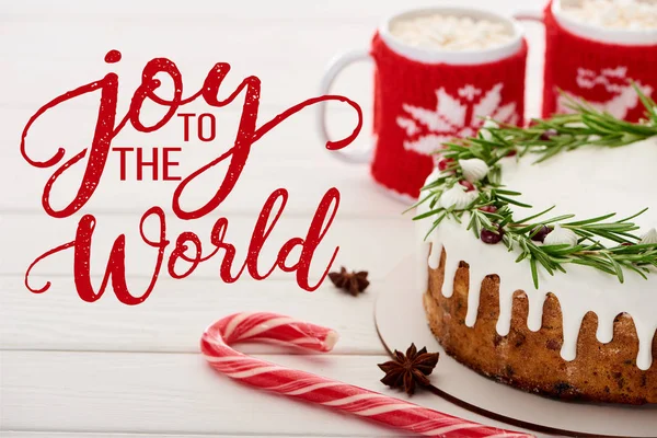 Candy cane, christmas pie with icing and two cups of cocoa with marshmallows on white wooden table with joy to the world illustration — Stock Photo