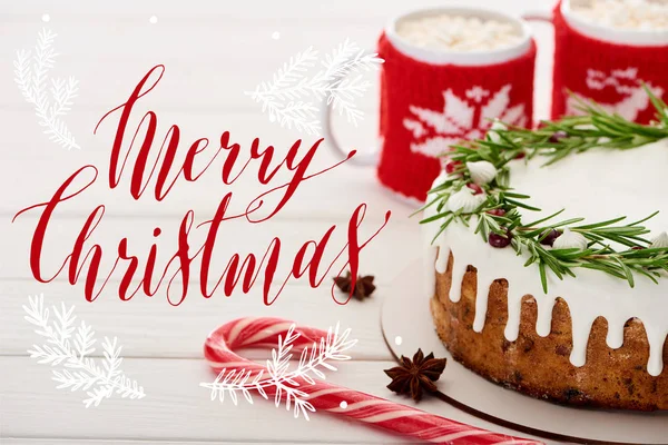 Bastoncino di zucchero, torta di Natale con glassa e due tazze di cacao con marshmallow sul tavolo di legno bianco con illustrazione Buon Natale — Foto stock
