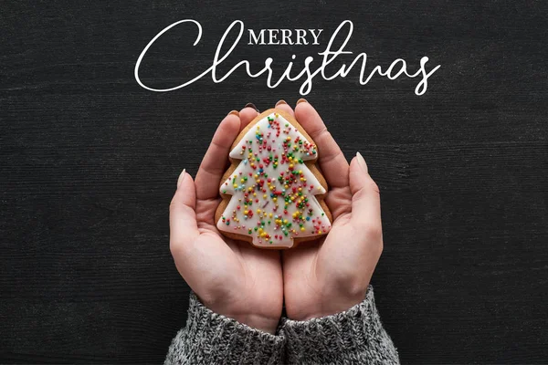 Cropped view of woman holding baked Christmas tree cookie in hands with Merry Christmas illustration — стокове фото