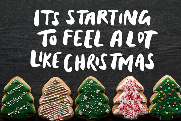 Flat lay with delicious glazed Christmas tree cookies on black background with its starting to feel a lot like Christmas illustration — Stock Photo