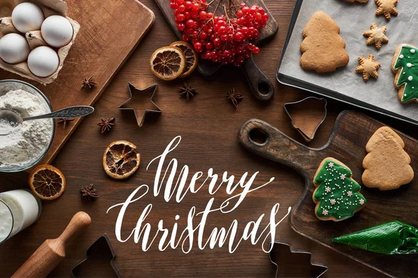 Vue du dessus des biscuits de Noël glacés près des ingrédients, des moules à pâte et du viorne sur une table en bois avec une illustration de Joyeux Noël — Photo de stock