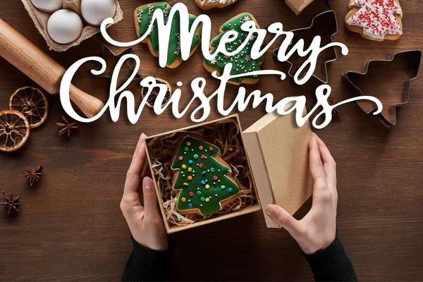 Vista cortada da caixa de presente de fechamento da mulher com biscoito de árvore de Natal na mesa de madeira com ilustração de Feliz Natal — Fotografia de Stock