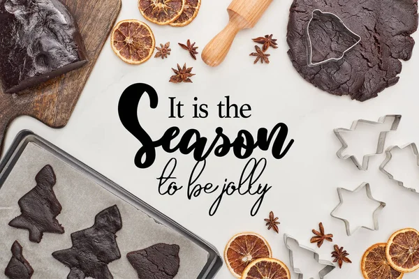 Vue du dessus de la pâte crue pour les biscuits de Noël au chocolat près des ingrédients sur fond blanc avec elle est la saison pour abeille lettrage joyeux — Photo de stock