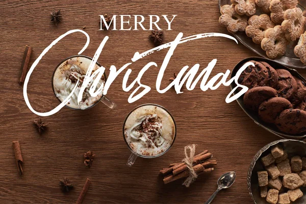 Top view of Christmas cacao with whipped cream on wooden table with spices and cookies with merry Christmas lettering — Stock Photo