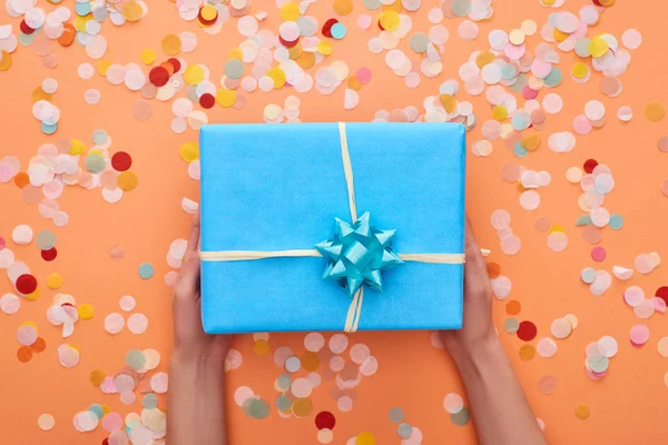 Cropped view of woman holding blue present with bow near confetti on orange — Stock Photo