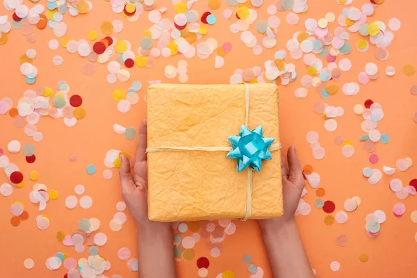 Cropped view of woman holding present with blue bow near confetti on orange — Stock Photo