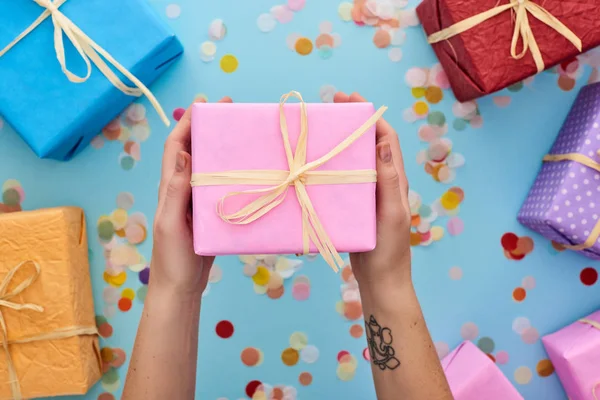 Vue recadrée d'une femme tenant une boîte cadeau rose enveloppée près de cadeaux colorés près de confettis sur bleu — Photo de stock