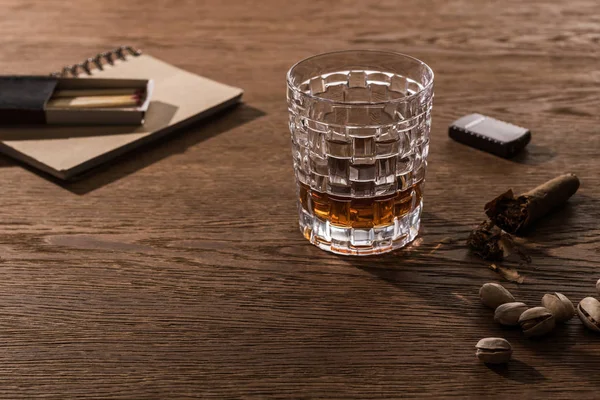 Brandy in glass with cigar, lighter and pistachios on wooden table — Stock Photo