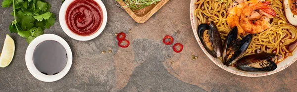 Top view of spicy seafood ramen near fresh ingredients and sauces on stone surface, panoramic shot — Stock Photo