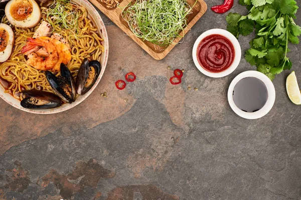 Vue de dessus des ramen épicés de fruits de mer près des ingrédients frais et des sauces sur la surface de pierre — Photo de stock