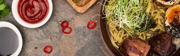 Top view of spicy meat ramen near fresh ingredients and sauces on stone surface, panoramic shot — Stock Photo