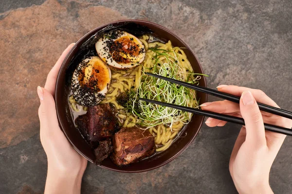 Cropped view of woman eating spicy meat ramen with chopsticks on stone surface — Stock Photo