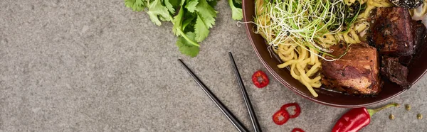 Top view of spicy meat ramen near chili pepper, parsley and chopsticks on grey concrete surface, panoramic shot — Stock Photo