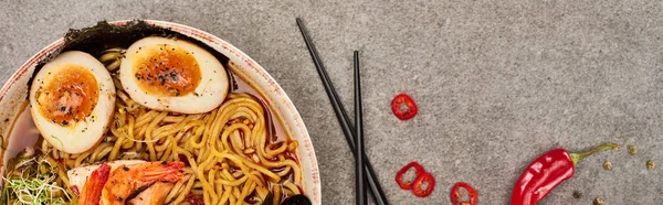 Draufsicht auf würzige Meeresfrüchte-Ramen in der Nähe von Chilipfeffer und Essstäbchen auf grauer Betonoberfläche, Panoramaaufnahme — Stockfoto