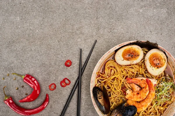 Top view of spicy seafood ramen near chili pepper and chopsticks on grey concrete surface — Stock Photo