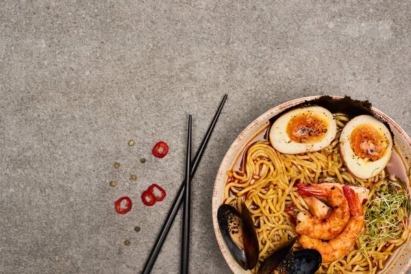 Top view of spicy seafood ramen near chili pepper and chopsticks on grey concrete surface — Stock Photo