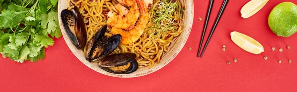 Blick von oben auf würzige Meeresfrüchte-Ramen in der Nähe von Essstäbchen, Petersilie und Limette isoliert auf rotem, Panoramaaufnahme — Stockfoto