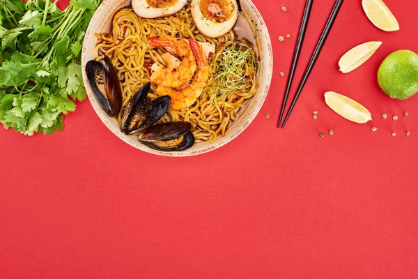 Top view of spicy seafood ramen near chopsticks, parsley and lime isolated on red — Stock Photo