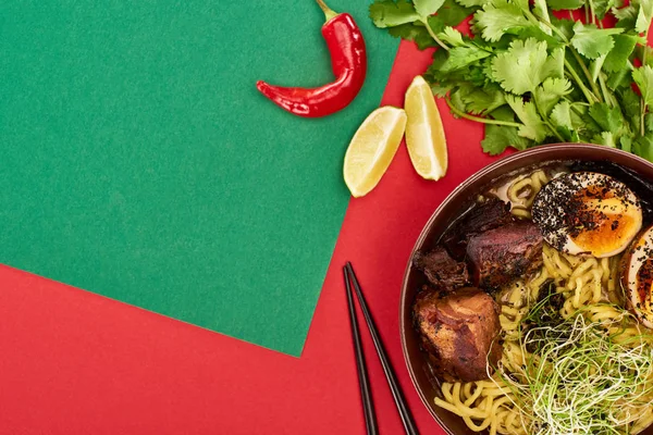 Vista superior de ramen carne picante perto de ingredientes frescos e pauzinhos na superfície verde e vermelha — Fotografia de Stock