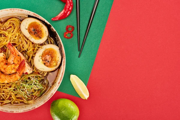 Vue de dessus des ramen épicés de fruits de mer près des ingrédients frais et des baguettes sur la surface verte et rouge — Photo de stock