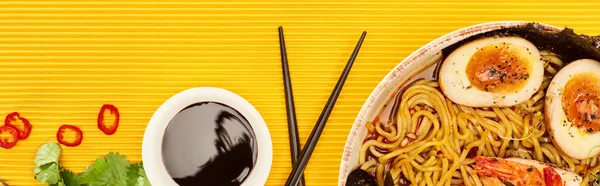 Top view of seafood ramen near fresh ingredients, soy sauce and chopsticks on yellow surface, panoramic shot — Stock Photo