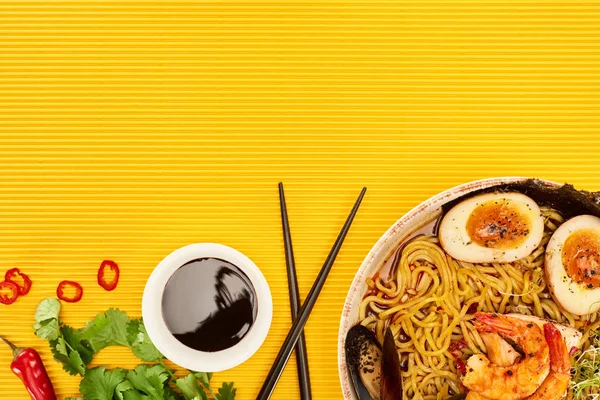 Top view of seafood ramen near fresh ingredients, soy sauce and chopsticks on yellow surface — Stock Photo