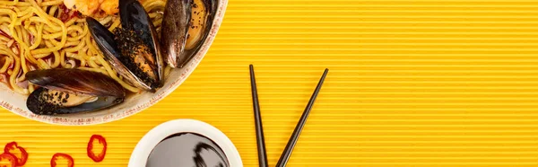 Top view of seafood ramen near soy sauce and chopsticks on yellow surface, panoramic shot — Stock Photo